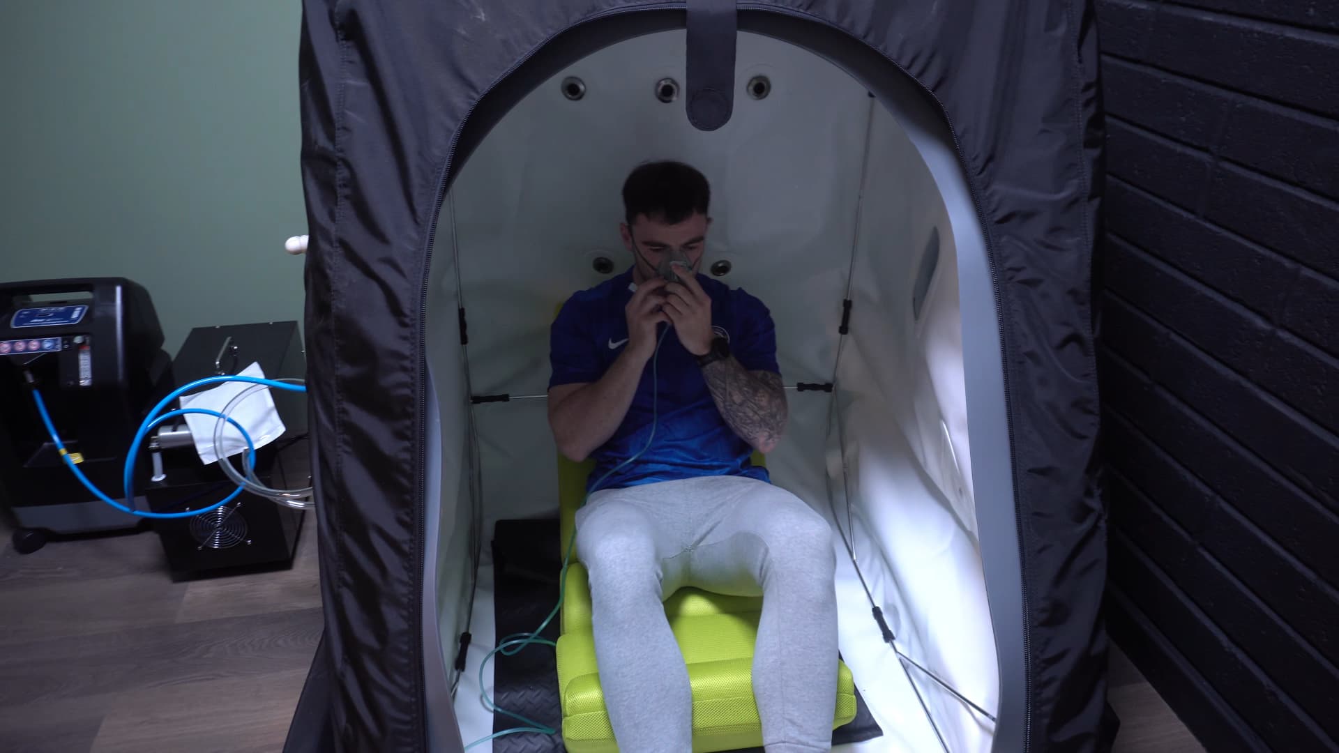 A client seated in the open hyperbaric chamber holding the oxygen mask, with the concentrator alongside